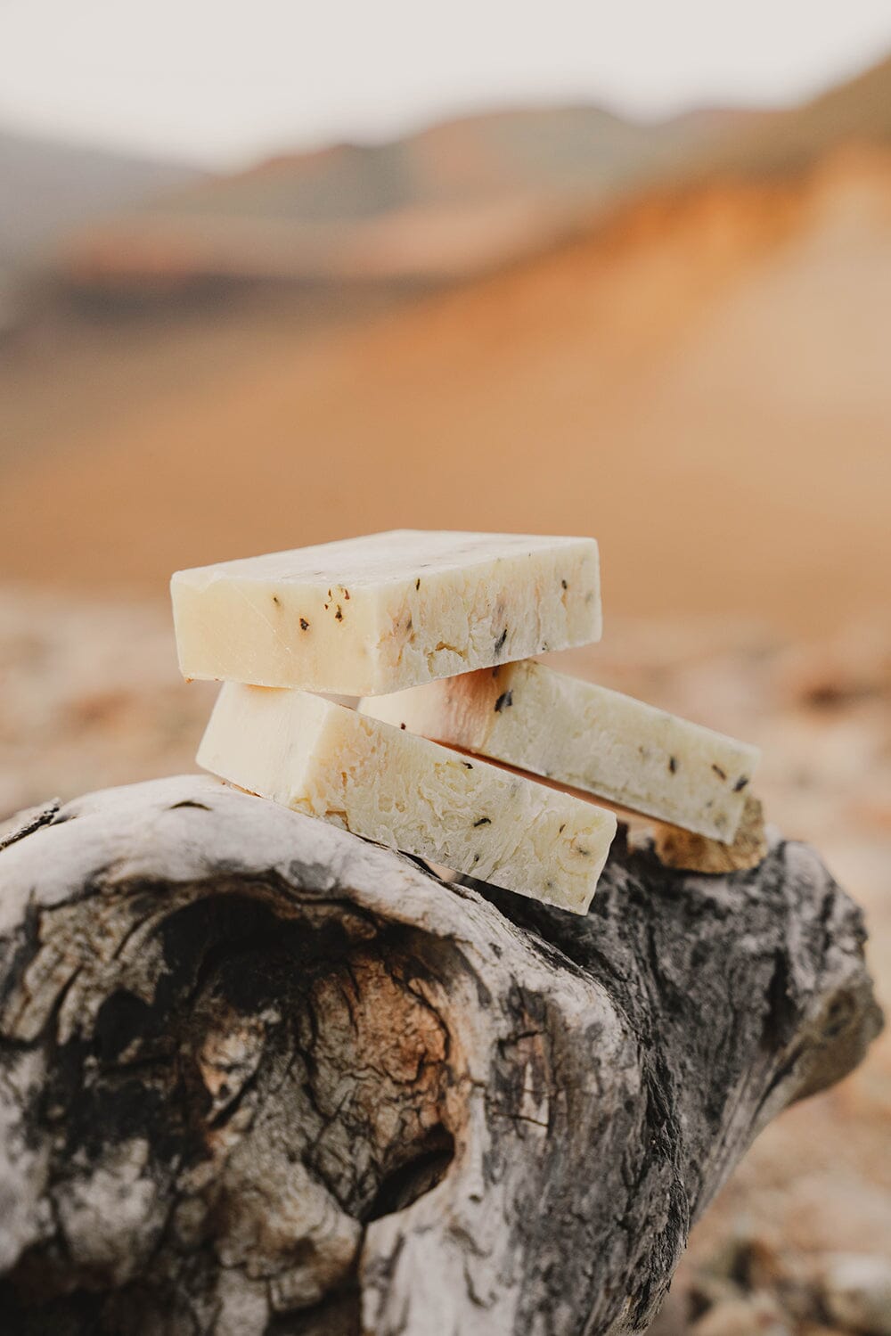 Bars of all-natural soap made with essential oils, such as eucalyptus, stacked on a log overlooking a sun kissed mountain range.