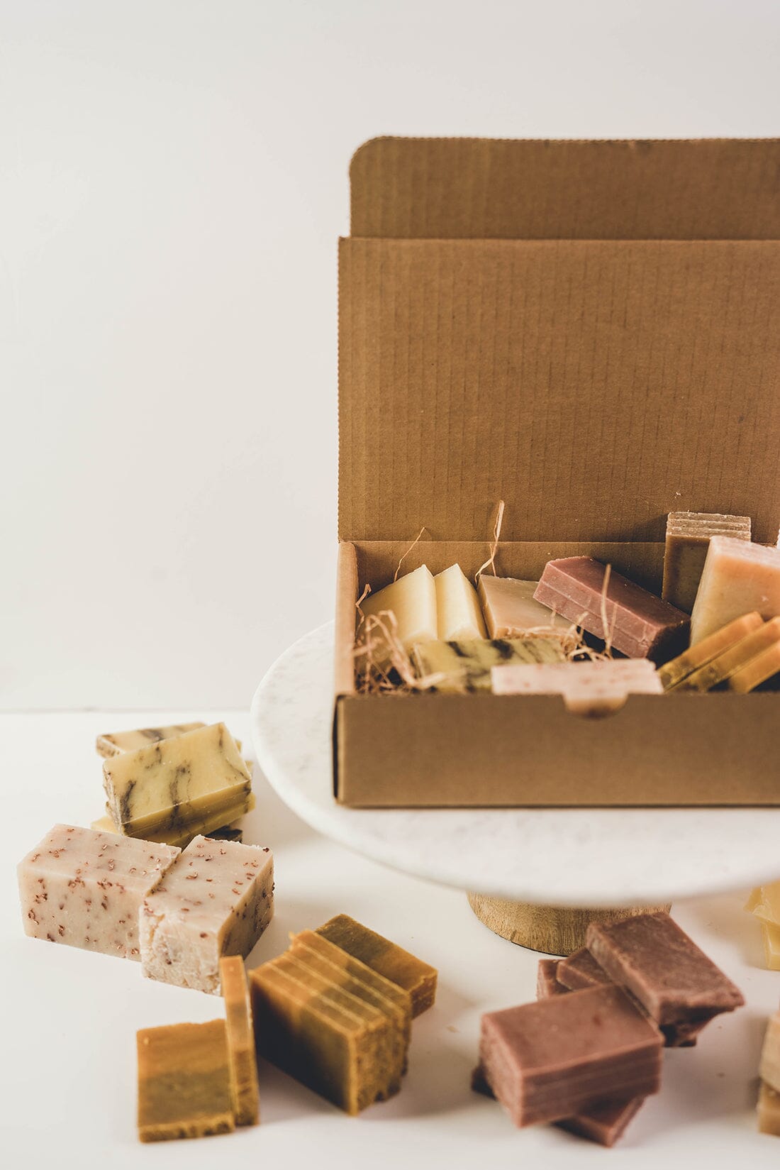  A cardboard box resting on a table that is packed with various samples of all natural bars of soap set on a white background.