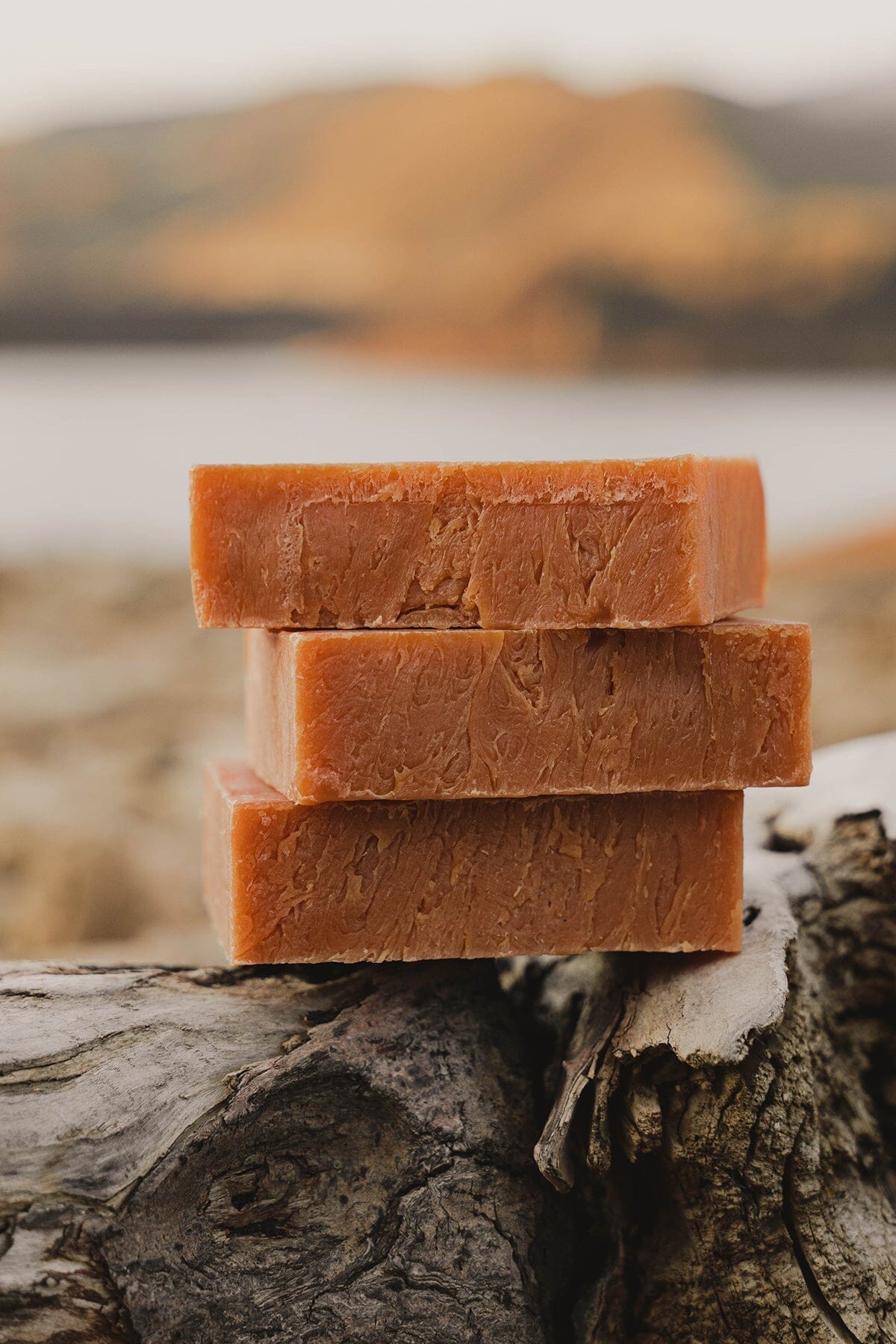 A stack of all-natural bars of soap made with dragon's blood sitting on top of a log overlooking a sun kissed lake set in a desert valley.