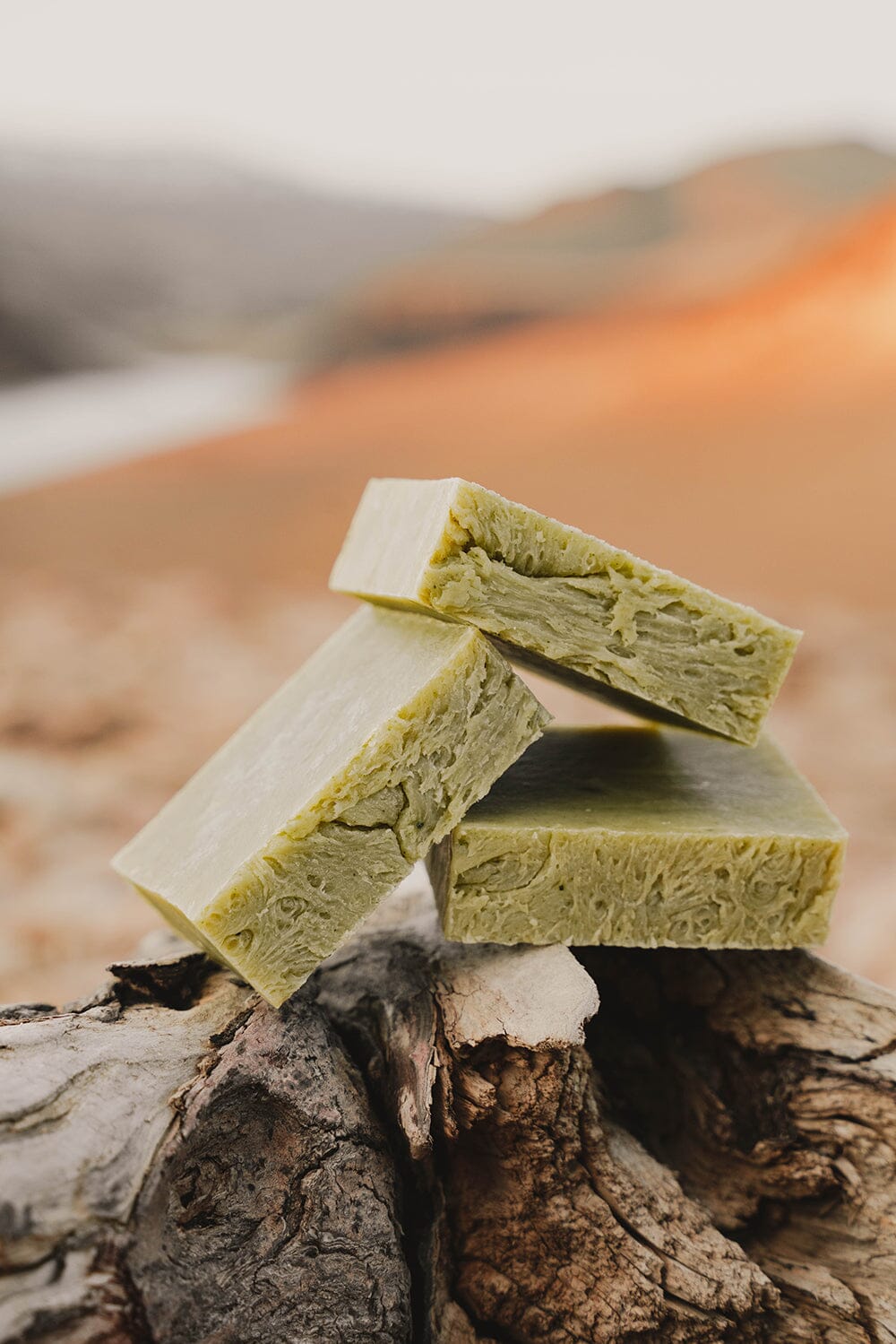 Bars of all-natural soap made with essential oils,  like peppermint and eucalyptus, stacked on a log overlooking a sun kissed valley with a lake in the distance.